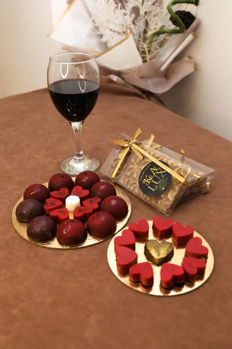 Praline i čokoladna srca na zlatnim tacnama i čokoladna tabla u poklon pakovanju, uz čašu crnog vina